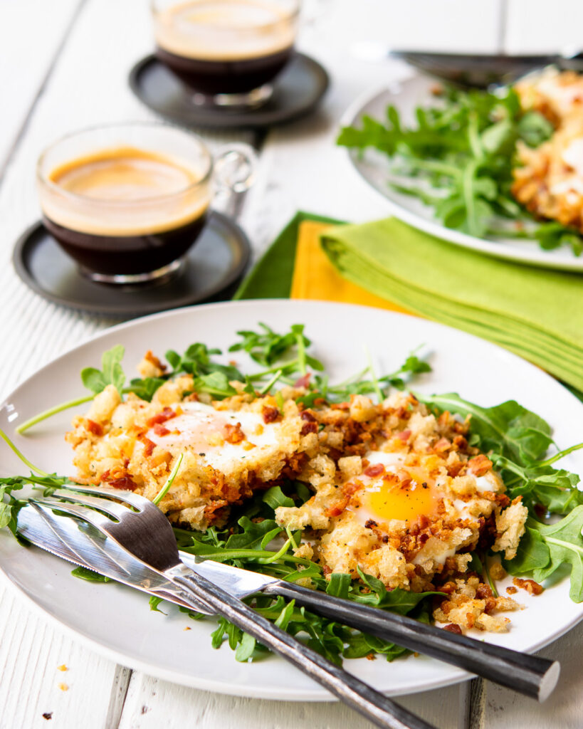 Two white plates with crispy eggs in bacon breadcrumbs on a bed of arugula with napkins and coffees.