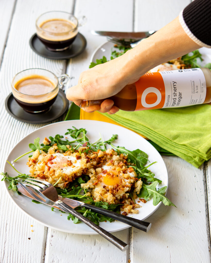 A hand sprinkling sherry vinegar over the top of crispy eggs in bacon breadcrumbs.