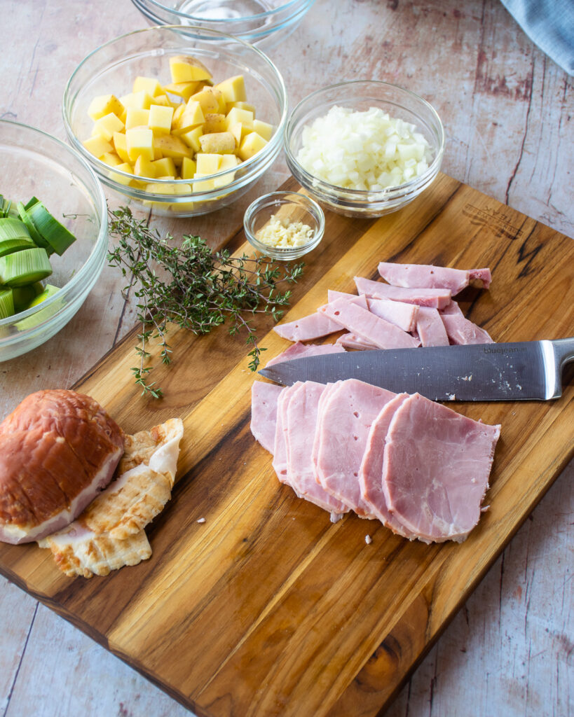 A cutting board with a knife cutting hamd into slices, and other ingredients around.