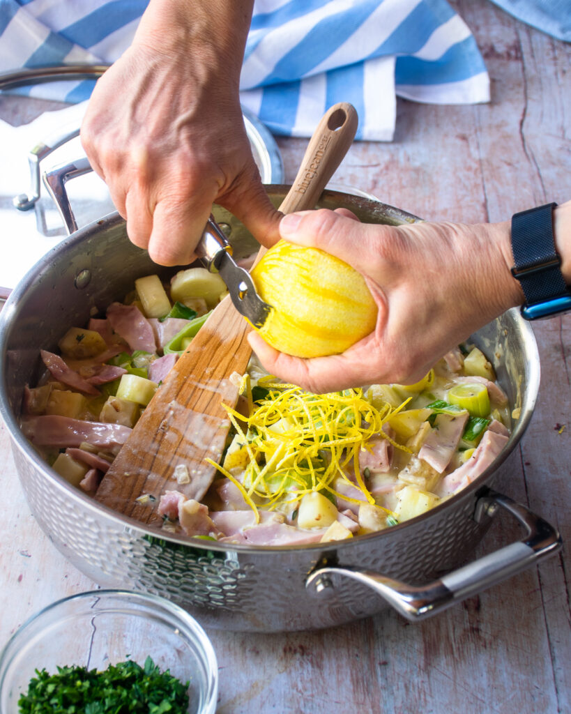 Hands zesting a lemon into a saucepan with ham, leek and potato pie filling inside.