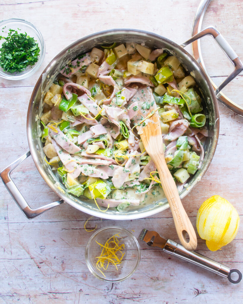 Ham leek and potato pie filling in a saute pan with a wooden spoon sticking out of it.