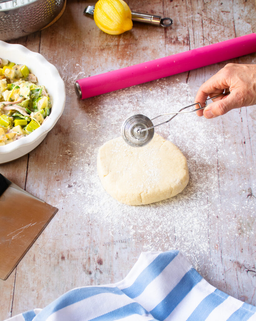 A hand spreading flour with a flour duster over pastry on a table with a rolling pin and kitchen towel near by.
