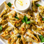 A plate of crispy artichokes on a white plate with a dip, lemon zest and parsley leaves.