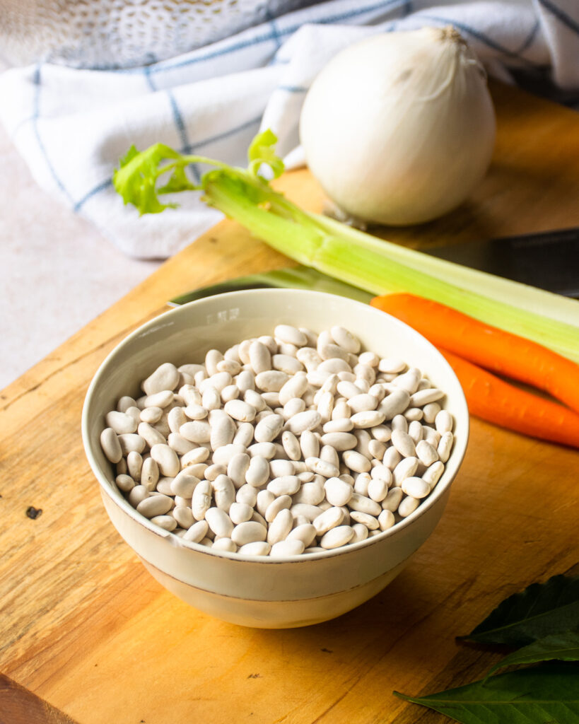 A bowl of white beans on a cutting board with vegetables behind it.