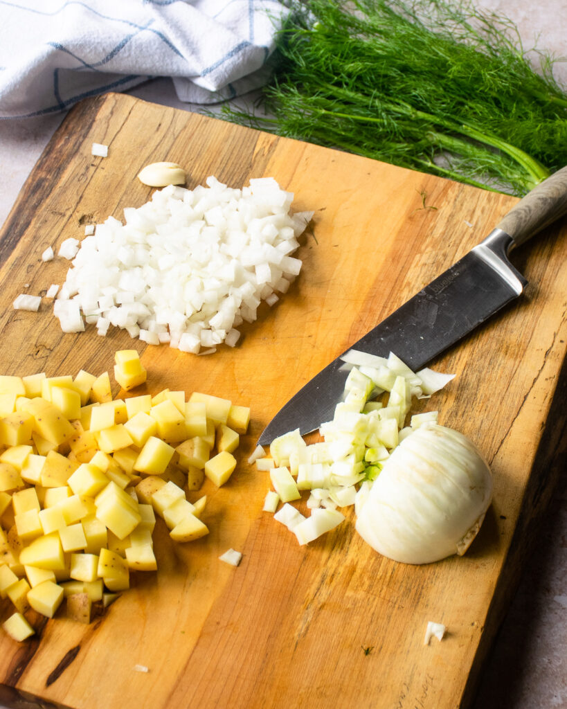 Fennel, onion and potato being chopped on a cutting board with some fresh dill in the background.