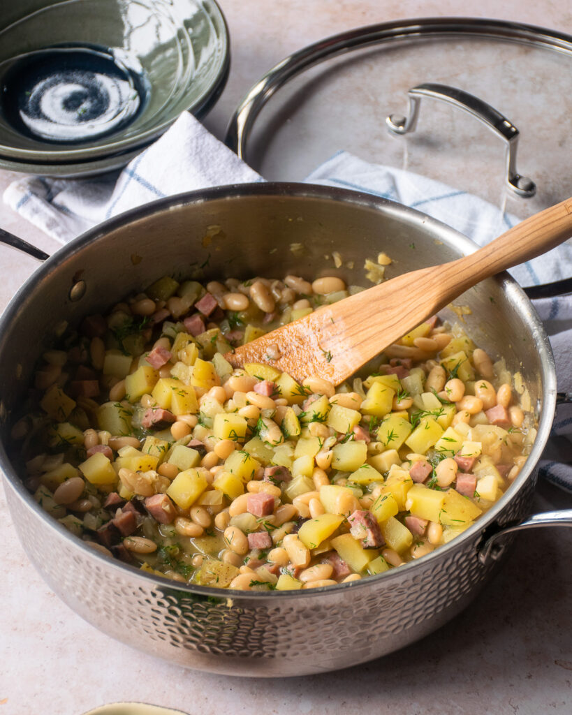 A hammered stainless steel saute pan with white beans and ham inside and a wooden spoon sticking out.