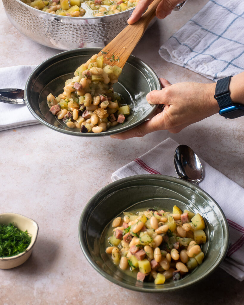 Hands serving white beans and ham into green ceramic bowls.