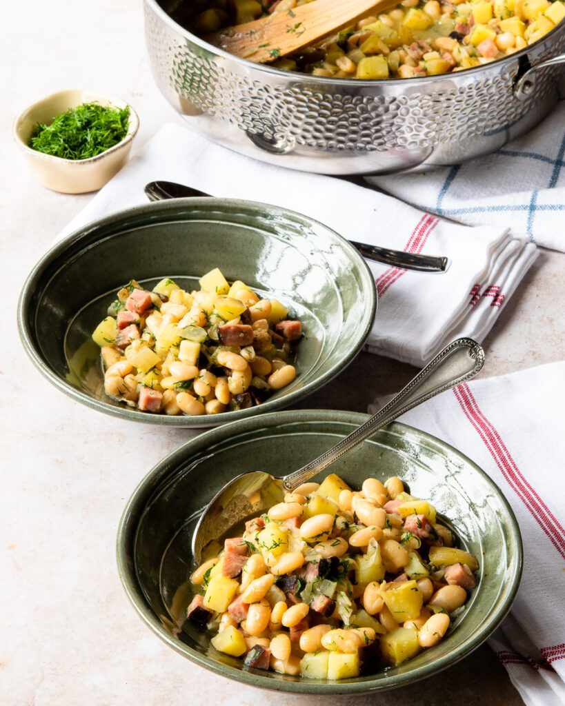 Two green bowls with white beans and ham inside with a hammered stainless steel sauté pan in the back ground.