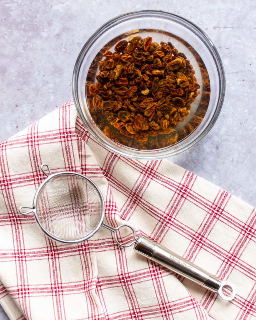 A bowl of raising plumping in hot water in a bowl with a red checked kitchen towel and a fine strainer.