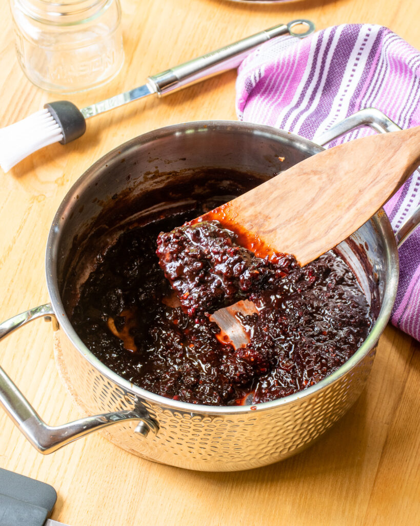 Blackberry BBQ sauce in a stainless steel saucepan with a wooden spoon lifting out of the pan.