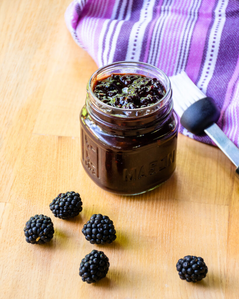 A small jar of blackberry BBQ sauce on a countertop with a purple kitchen towel, a basting brush and a few blackberries.