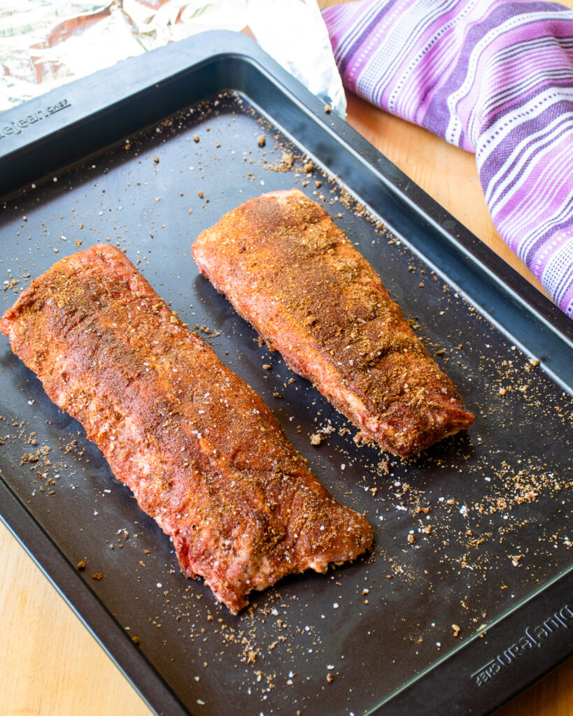 Two racks of spice-rubbed baby back ribs on a baking sheet.
