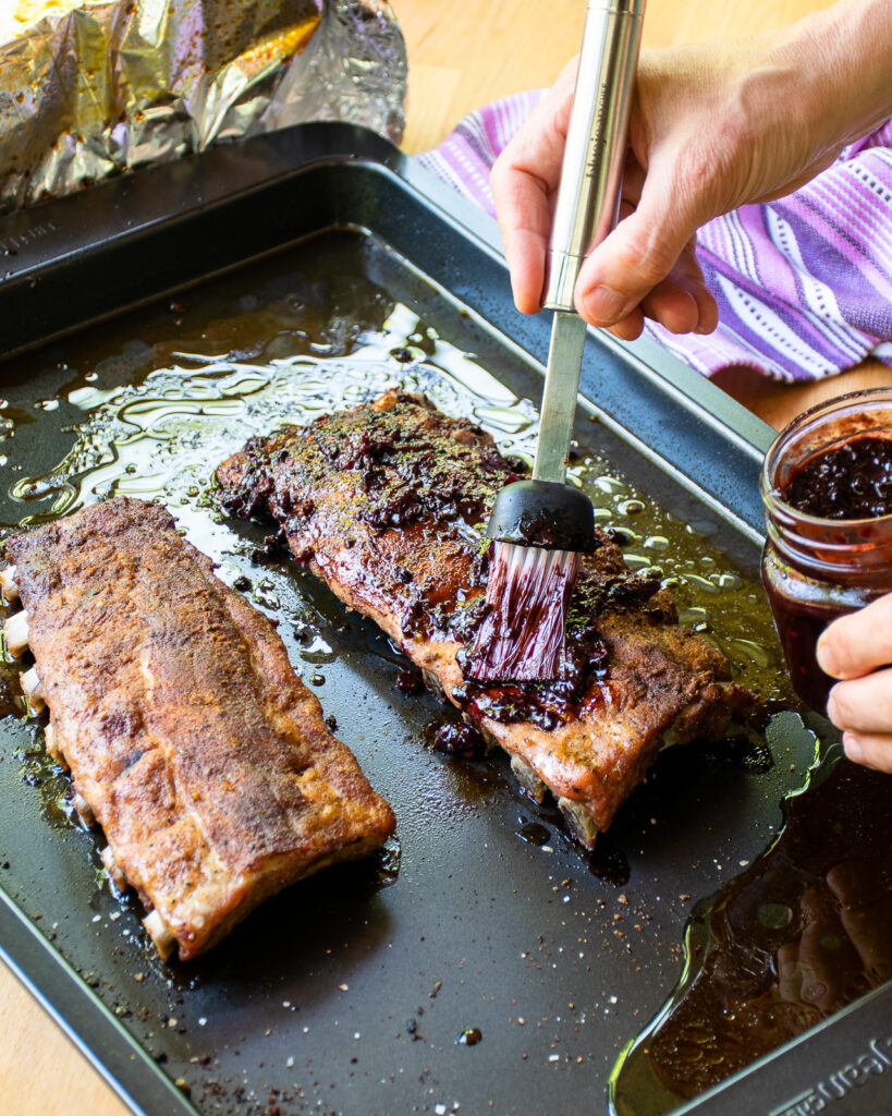 Hands brushing blackberry BBQ sauce onto a cooked rack of ribs.