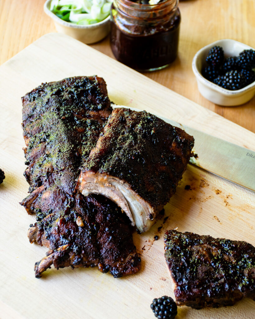 Two racks of baby back ribs with blackberry BBQ sauce on a cutting board - one of the racks cut in half.