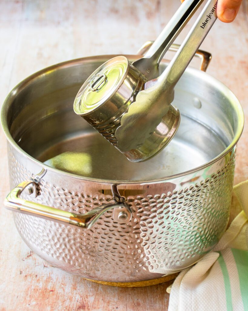 Tongs lifting a can out of a stainless steel pot of water.