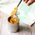 A spoon spooning dulce de leche out of a can with a kitchen towel in the background.