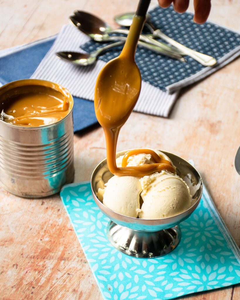 A spoon drizzling dulce de leche over a silver bowl of vanilla ice cream.