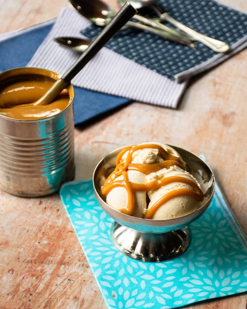 A silver ice cream bowl with vanilla ice cream and dulce de leche on a turqoise napkin with an open can of dulce de leche next to it.