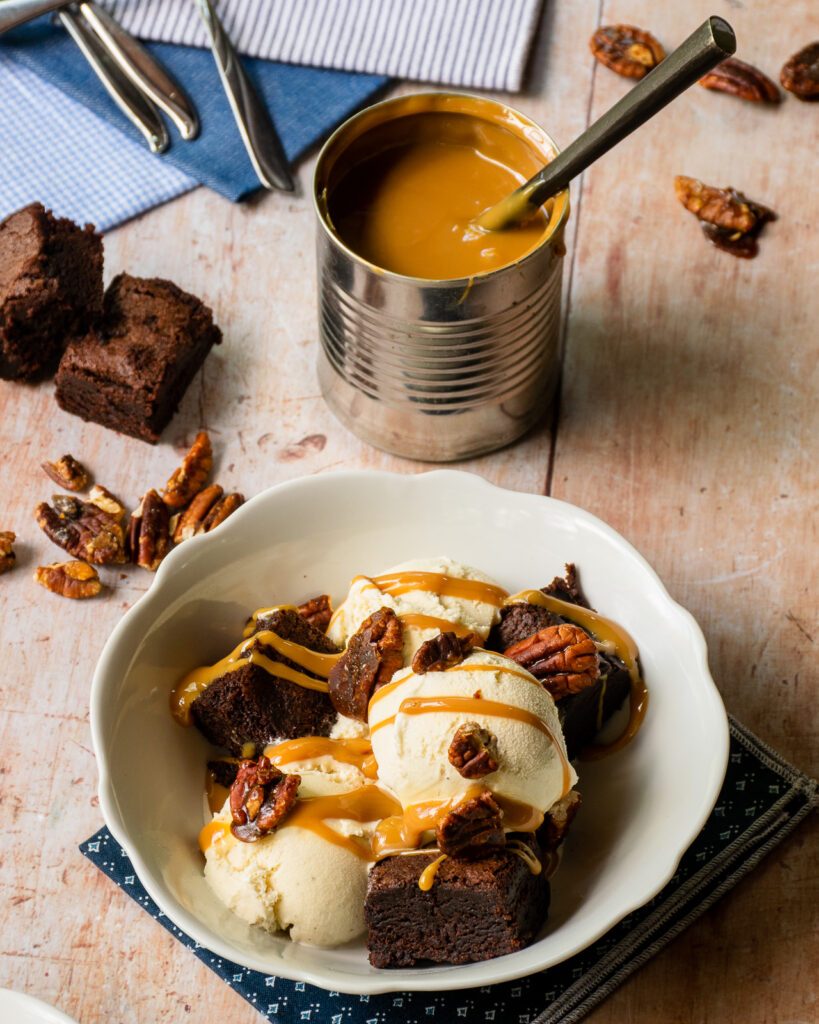 A white bowl of ice cream and brownies in front of a can of dulce de leche with a spoon sticking out of it.