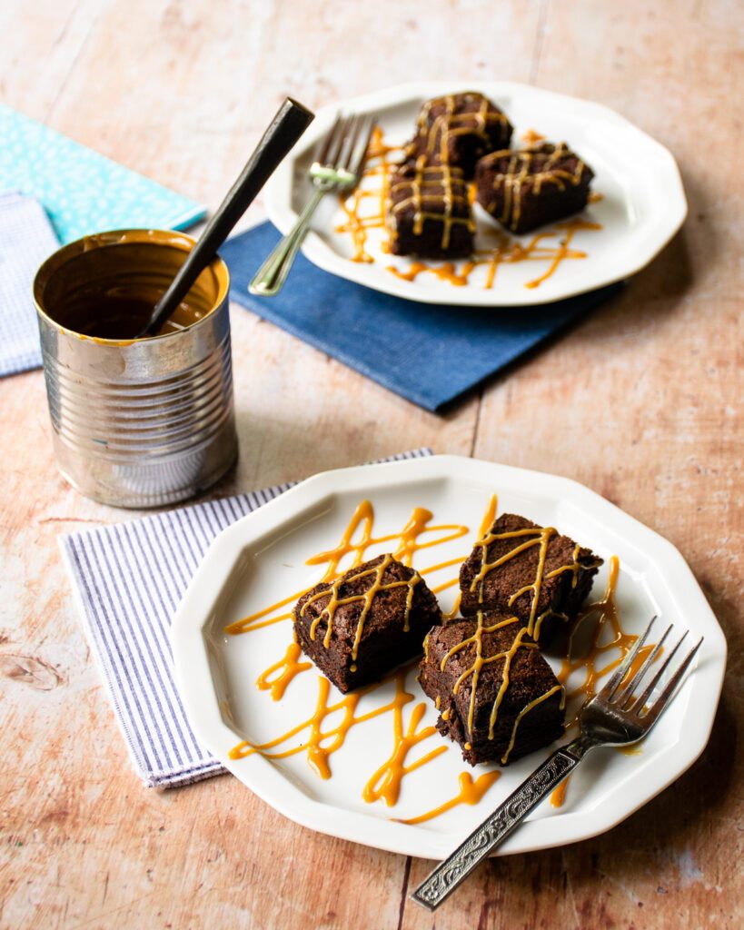 Two plates of brownies drizzled with dulce de leche with a can of dulce de leche next to them with a spoon sticking out of it.