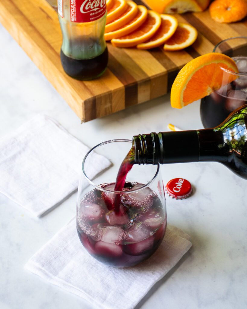 Wine being poured into a stemless wine glass with slices of orange, a jigger and cutting board near by.