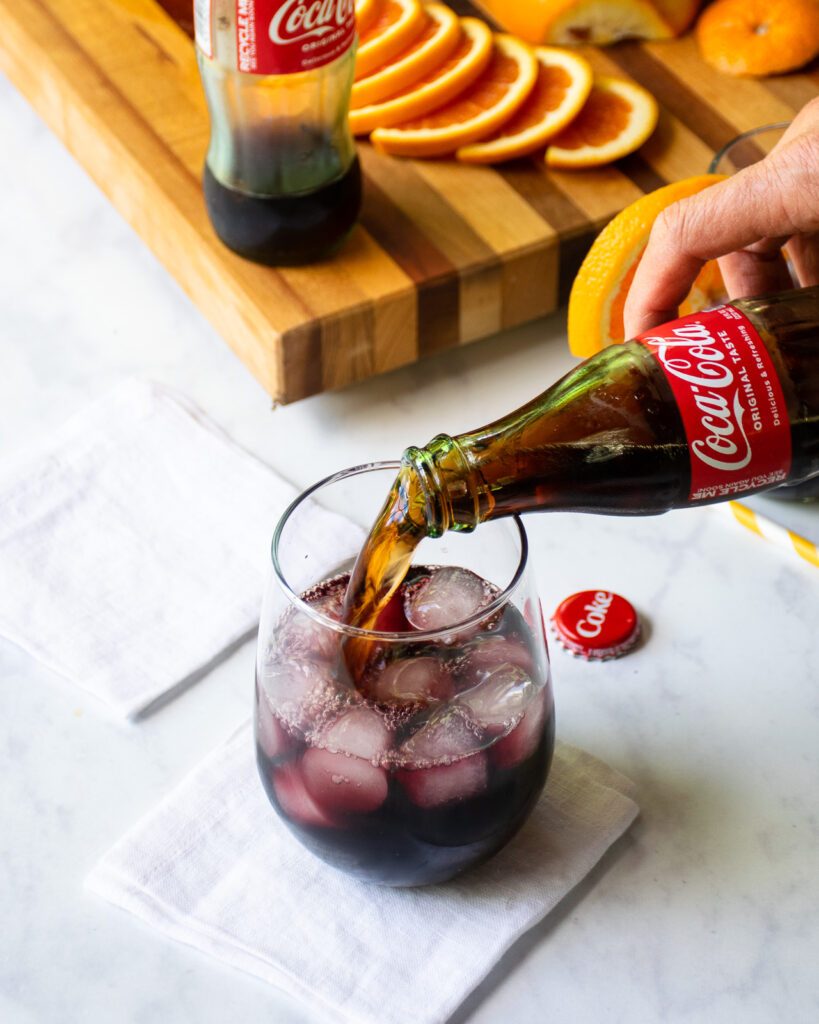 Topping a glass of red wine with coca-cola with orange slices and a jigger in the background.