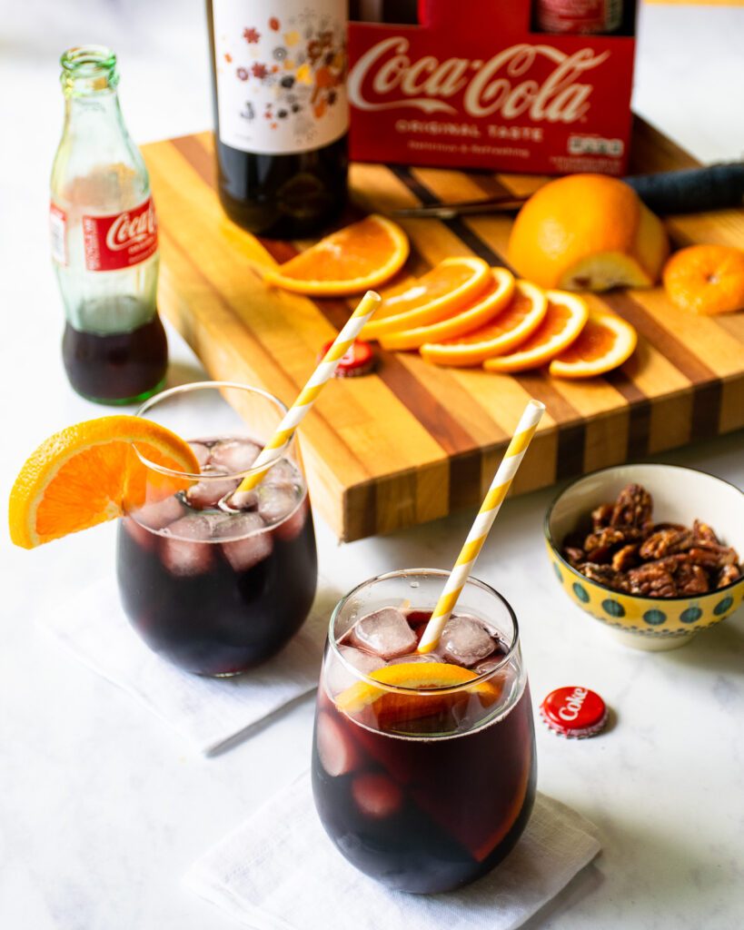 Two kalimoxto cocktails on a counter with a bottle of coca-cola, a bottle of red wine and some orange slices in the background.