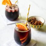 Two stemless wine glasses with Kalimoxto drinks and an orange slice inside, with a bowl of nuts nearby.