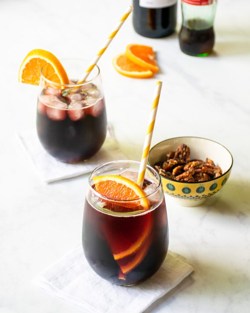 Two stemless wine glasses with Kalimoxto drinks and an orange slice inside, with a bowl of nuts nearby.