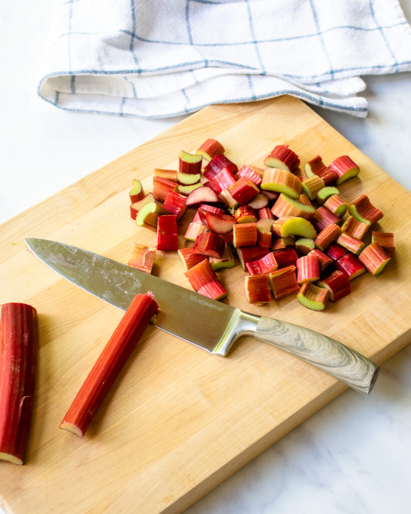 A knife cutting rhubarb stalks into chunks on a wooden cutting board.