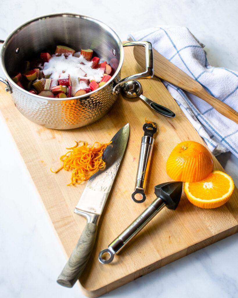 Ingredients for rhubarb fool on a cutting board, with sugar and rhubarb in a stainless steel saucepan, orange zest with a knife and zester and a halved orange with a reamer.