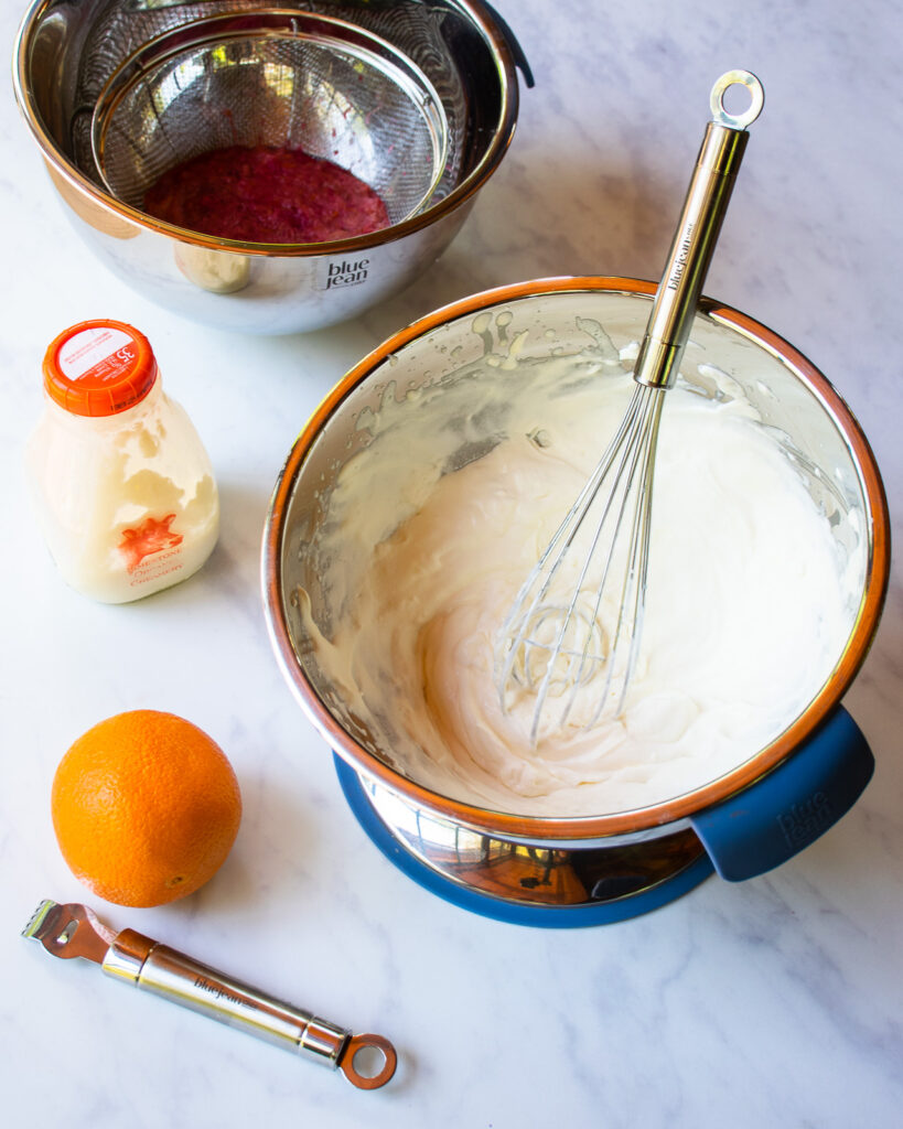 Whipped cream in a stainless steel bowl with an orange and zester near by.