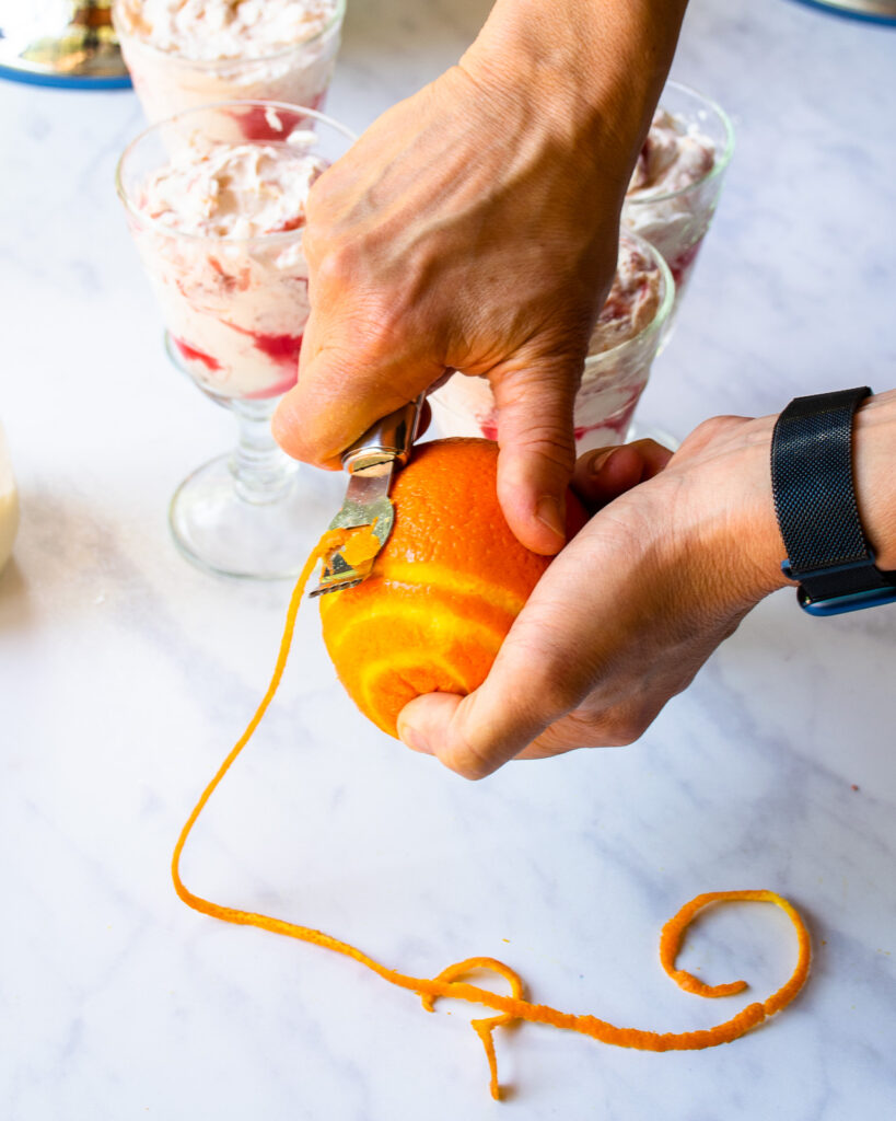 Hands using a channel knife to remove a long piece of orange zest from an orange.