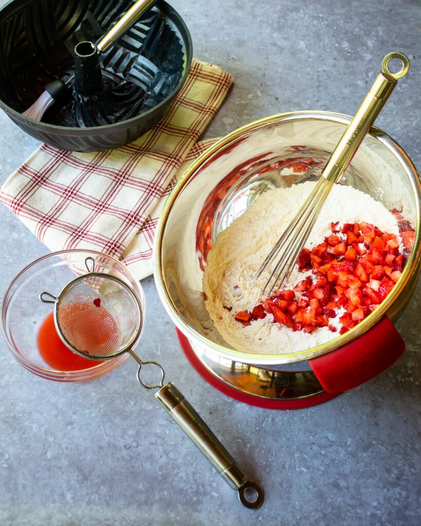 A stainless steel bowl with dry ingredients and diced strawberries inside, and a ball whisk sticking out.