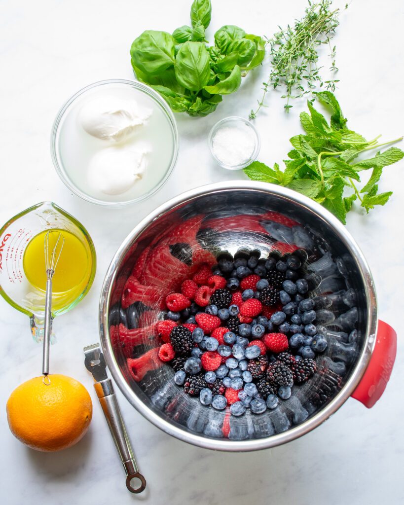 Ingredients for burrata and berry appetizer on a marble countertop - a bowl of berries, vinaigrette, an orange and zester, burrata and fresh herbs.