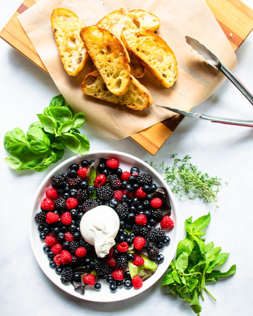 Looking down on burrata and berry appetizer in progress - a plate of berries with burrata in the center and some fried bread and tongs nearby.