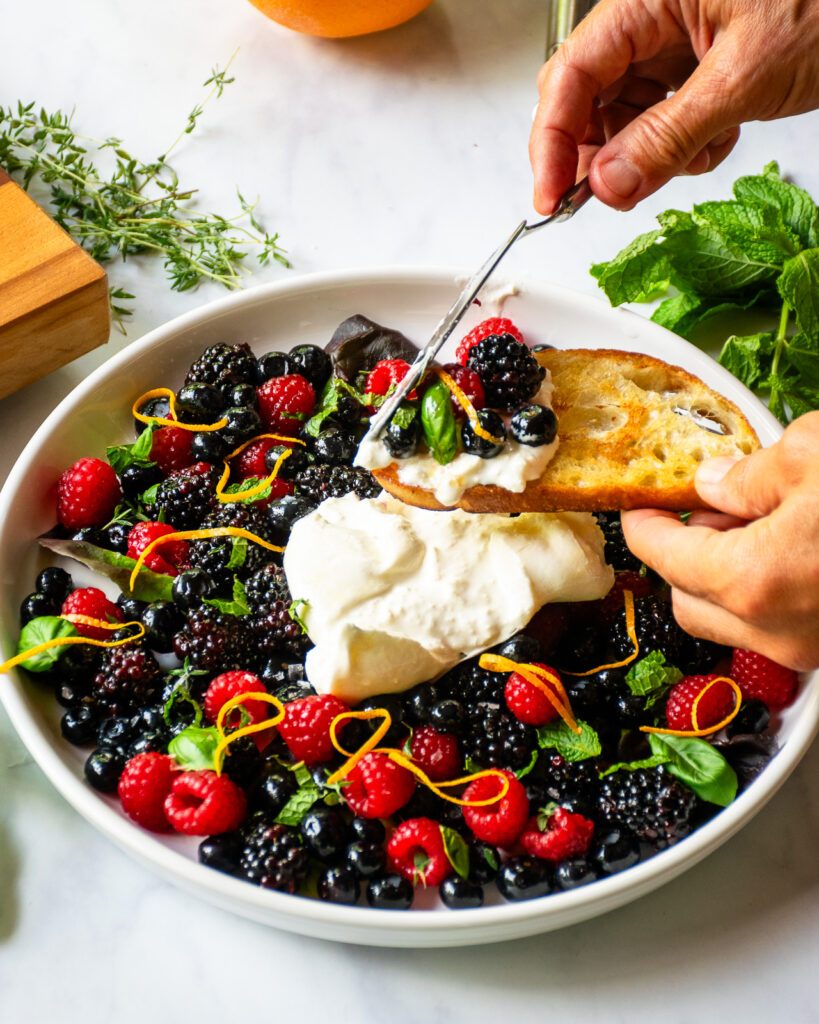 Hands spreading some burrata and berries onto a ciabatta crostini.