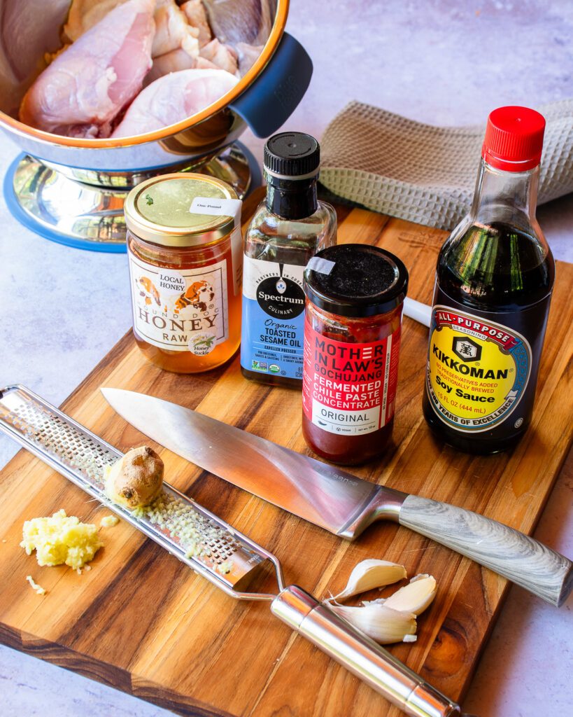 Ingredients for a marinade for korean grilled chicken on a cutting board - grated ginger, garlic, gochujang, soy sauce, honey, sesame oil and a bowl of raw chicken in the background.