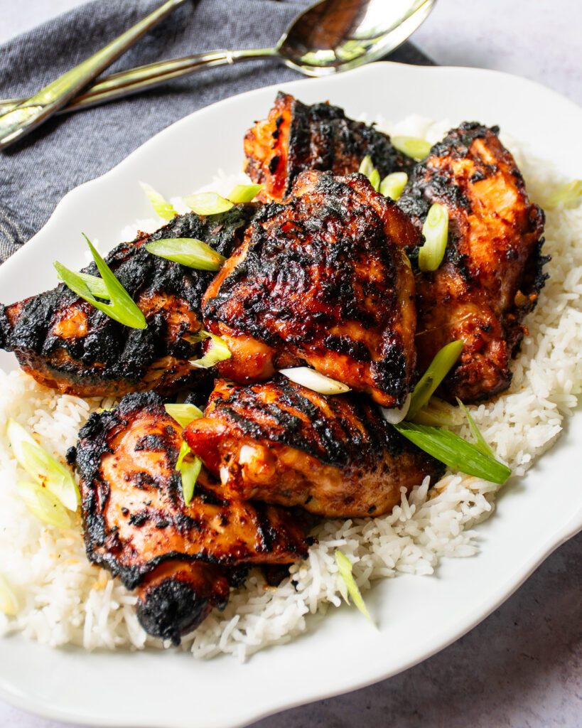 Looking down on a white oval platter with korean grilled chicken on top of a bed of rice, a grey napkin and serving utensils nearby.