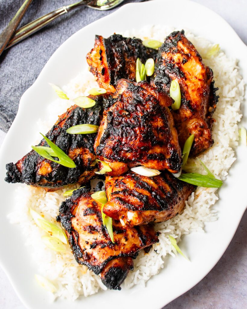 Looking down on a white oval platter with korean grilled chicken on top of a bed of rice, a grey napkin and serving utensils nearby.