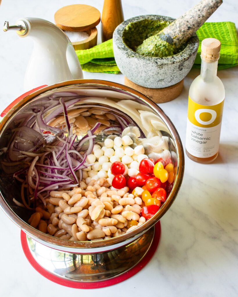 A bowl with white beans, tomatoes, fresh mozzarella and red onion in front of a mortar and pestle and a bottle of white balsamic vinegar.