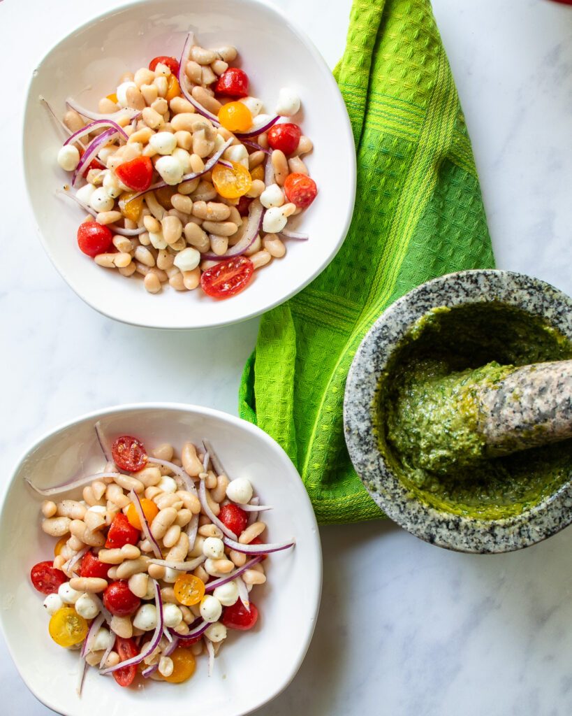 Looking down on two bowls of white bean salad with a mortar and pestle of basil pesto and a green kitchen towel.