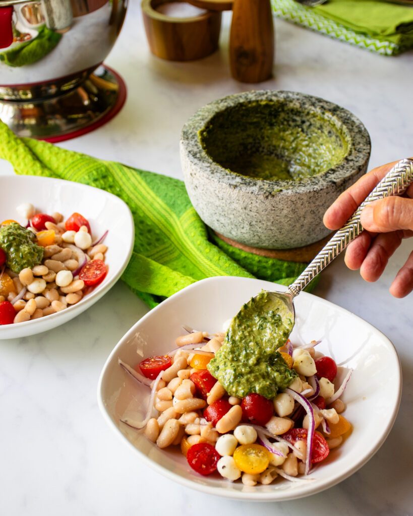 A hand dolloping pesto on a bowl of white beans with tomatoes and fresh mozzarella, with a mortar and pestle in the background.