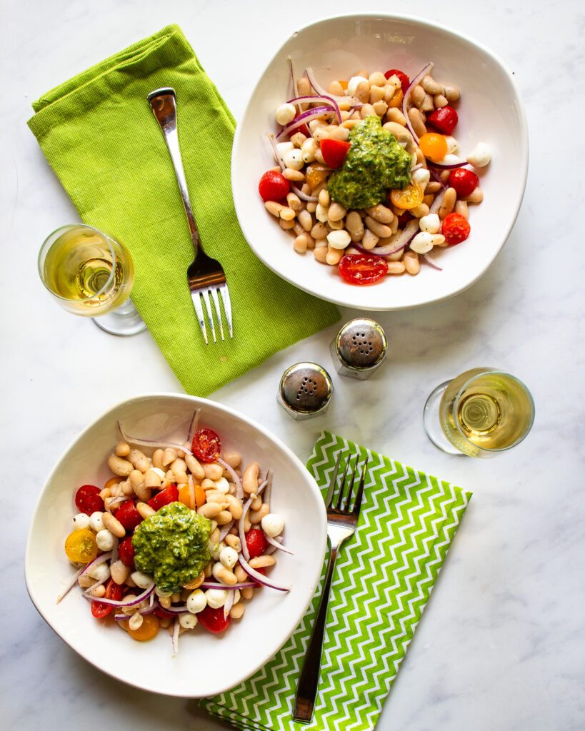 Two white bowls on a marble counter with pesto white beans, a green napkin, a fork, glass of white wine and salt and pepper shakers.