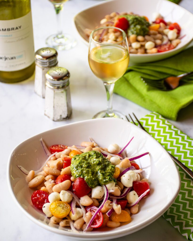 Two white bowls on a marble counter with pesto white beans, a green napkin, a fork, glass of white wine and salt and pepper shakers.