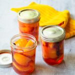 Three jars of pickled peaches, two with lids on and one with the lid off. A yellow kitchen towel in the background.