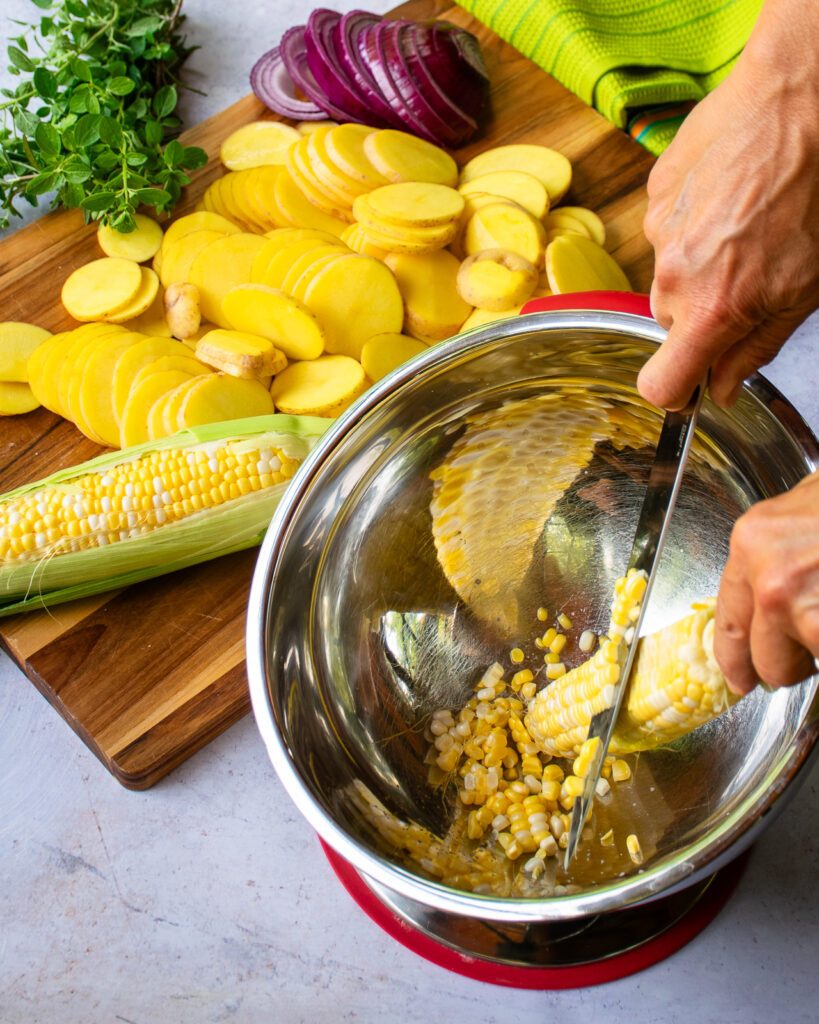 Hands cutting kernels off a cob of corn into a bowl with sliced potatoes and red onion and herbs in the background.