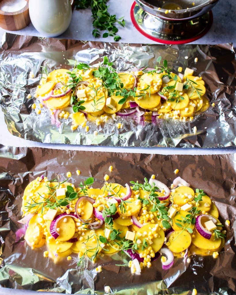 Two foil packet vegetable recipes being prepared - raw potatoes, corn, red onion, herbs on two pieces of aluminum foil