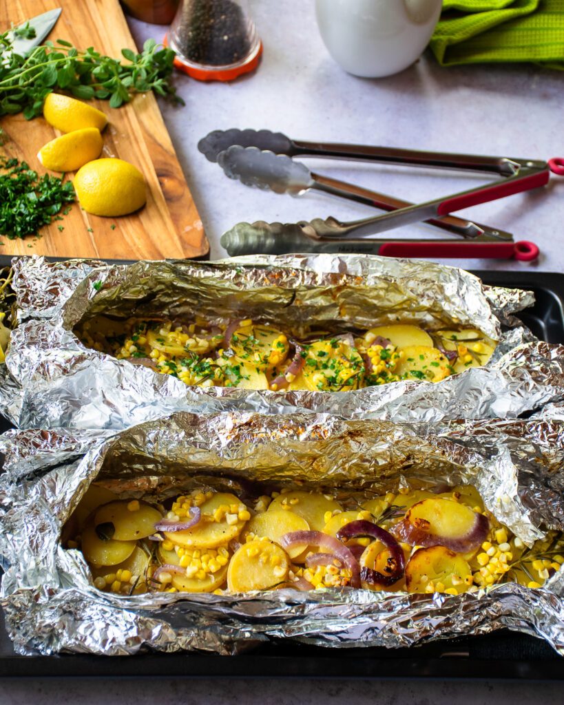 Two foil vegetable packets after being grilled on a countertop with tongs, lemon and fresh herbs in the background.