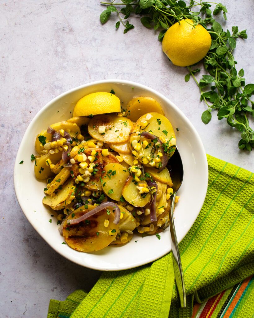 Corn, potatoes and red onion - vegetable hobo packets - in a white bowl with a green kitchen towel, herbs and half a lemon nearby.